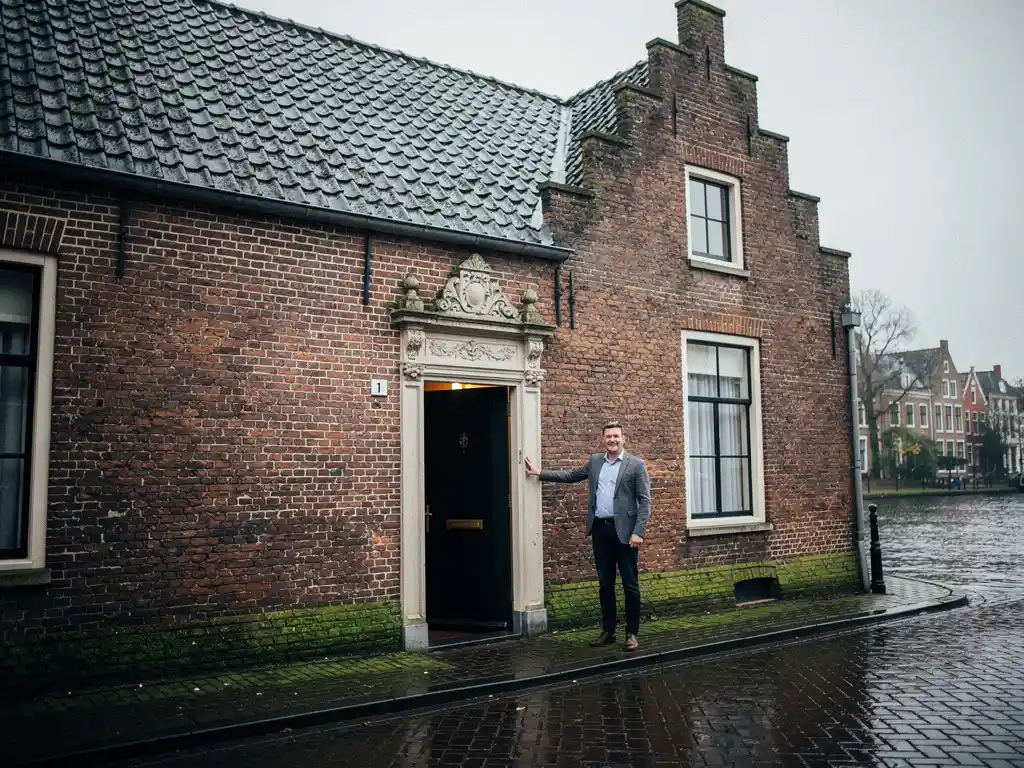 Makelaar in casual zakelijke kleding bij de voordeur van een 17e-eeuws Gouds grachtenpand met trapgevel en natte kinderkopjes.